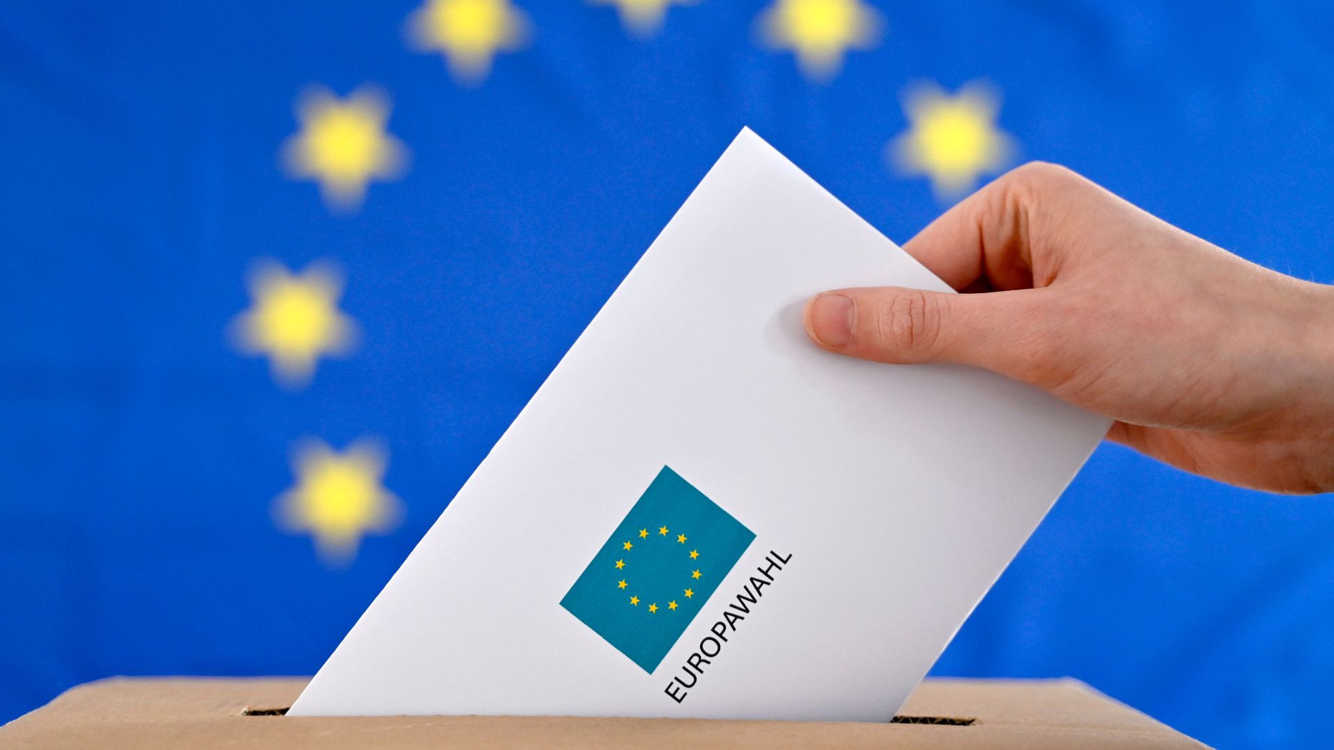 Eine Hand steckt einen Wahlbrief in eine Wahlurne. Auf dem Brief steht "Europawahl", im Hintergrund ist eine Europaflagge mit gelben Sternen auf blauem Hintergrund zu sehen.
