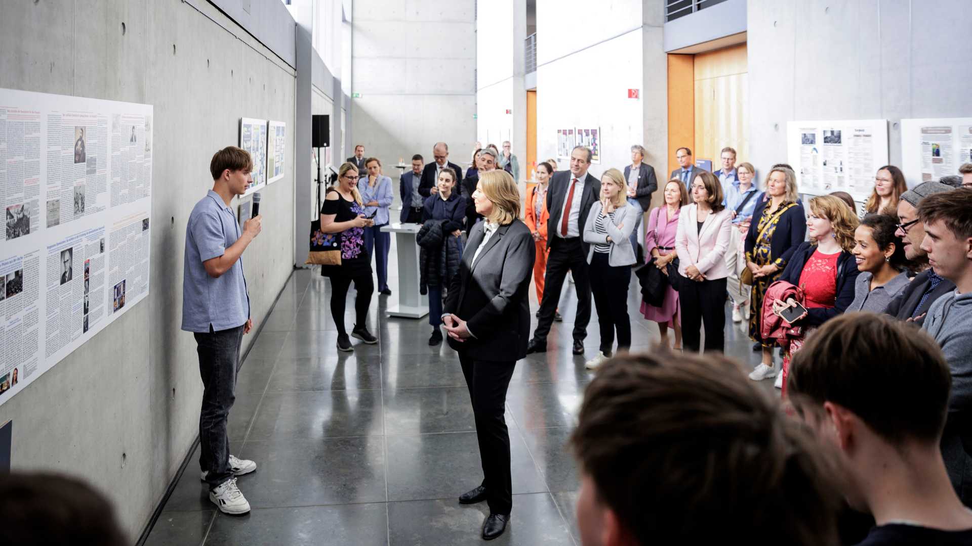 Leute stehen in einem Halbkreis und sehen einen Schüler an, der in ein Mikro spricht. Vor ihm steht Bundestagspräsidentin Bärbel Bas. Er erklärt die Arbeit, mit der er gemeinsam mit seinem Geschichtsleistungskurs am Schülerwettbewerb teilgenommen hat. Die Arbeit hängt hinter ihm an der Wand.